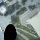 Aerial view from a high ledge showing a city square, roads, and a circular plaza, with dark shoes visible at the bottom edge as if standing on a tall structure.