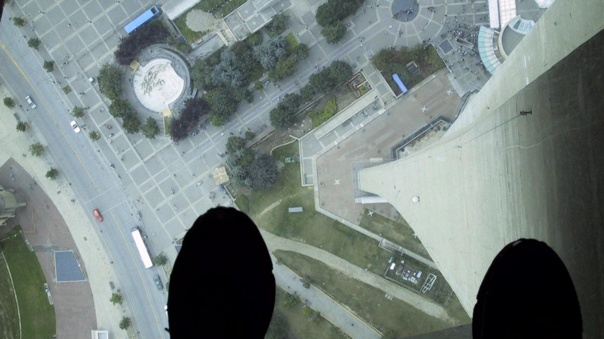 Aerial view from a high ledge showing a city square, roads, and a circular plaza, with dark shoes visible at the bottom edge as if standing on a tall structure.