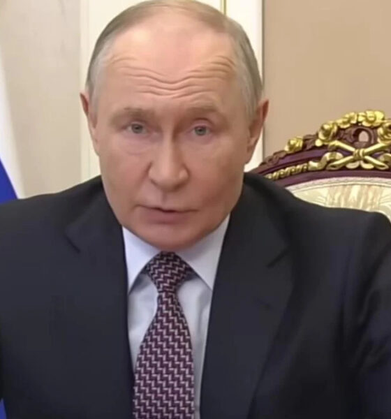 A man in a dark suit and patterned tie sits at a desk in a formal room with a decorative chair and a blue flag behind him, addressing an audience or meeting room.