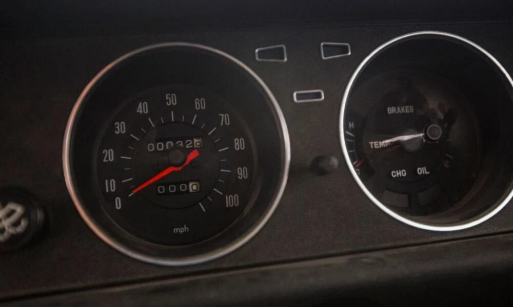 Close-up of a car dashboard with a speedometer and secondary gauges; the red needle sits near 0 mph, ready to drive.