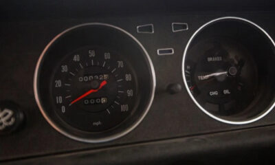 Close-up of a car dashboard with a speedometer and secondary gauges; the red needle sits near 0 mph, ready to drive.
