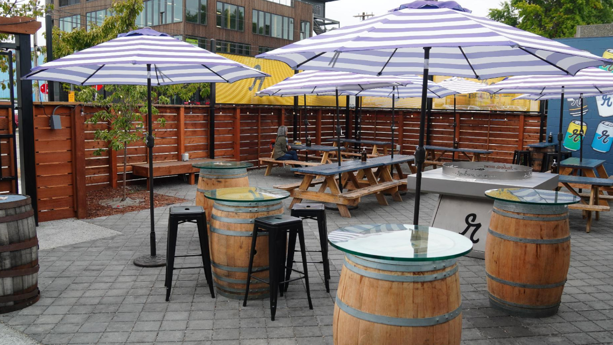Outdoor beer garden patio with striped purple-and-white umbrellas, wooden barrel tables, and picnic tables; a person sits at a table in the background.