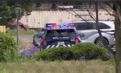 Seattle Police patrol car with red and blue lights on in a yard, parked behind a hedge and near an industrial backdrop.