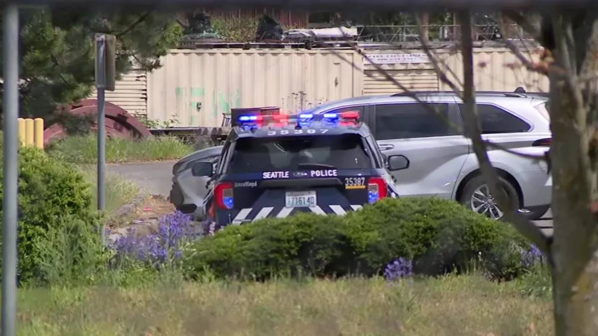 Seattle Police patrol car with red and blue lights on in a yard, parked behind a hedge and near an industrial backdrop.