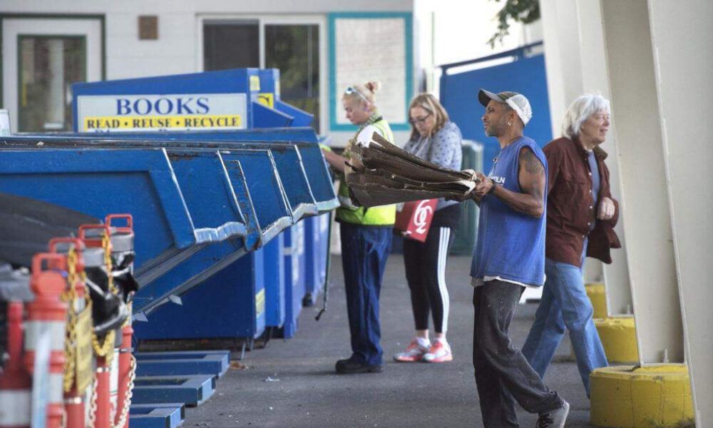 Man in a blue sleeveless shirt and cap carries flattened cardboard toward large blue recycling dumpsters outside a building, with bystanders nearby.