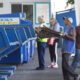 Man in a blue sleeveless shirt and cap carries flattened cardboard toward large blue recycling dumpsters outside a building, with bystanders nearby.