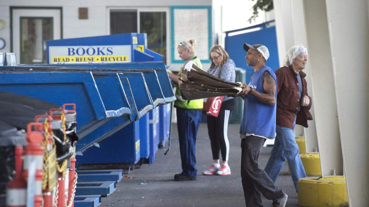 Man in a blue sleeveless shirt and cap carries flattened cardboard toward large blue recycling dumpsters outside a building, with bystanders nearby.