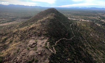 Aerial view of a hill with ancient circular earthworks at its base, set in a desert landscape with distant mountains and a trail winding up the slope (INAH watermark).