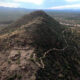 Aerial view of a hill with ancient circular earthworks at its base, set in a desert landscape with distant mountains and a trail winding up the slope (INAH watermark).