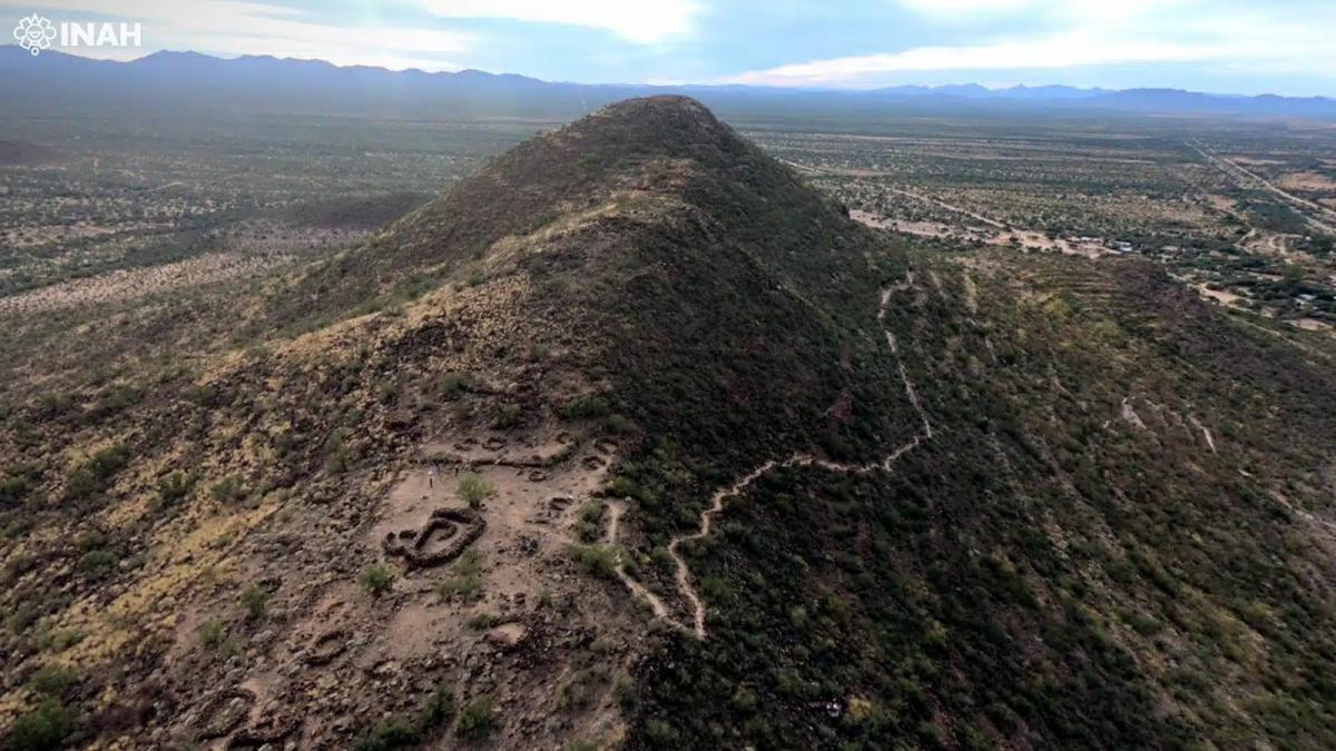 Aerial view of a hill with ancient circular earthworks at its base, set in a desert landscape with distant mountains and a trail winding up the slope (INAH watermark).