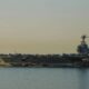 An aircraft carrier on calm water with numerous planes arranged on its deck at sunset.