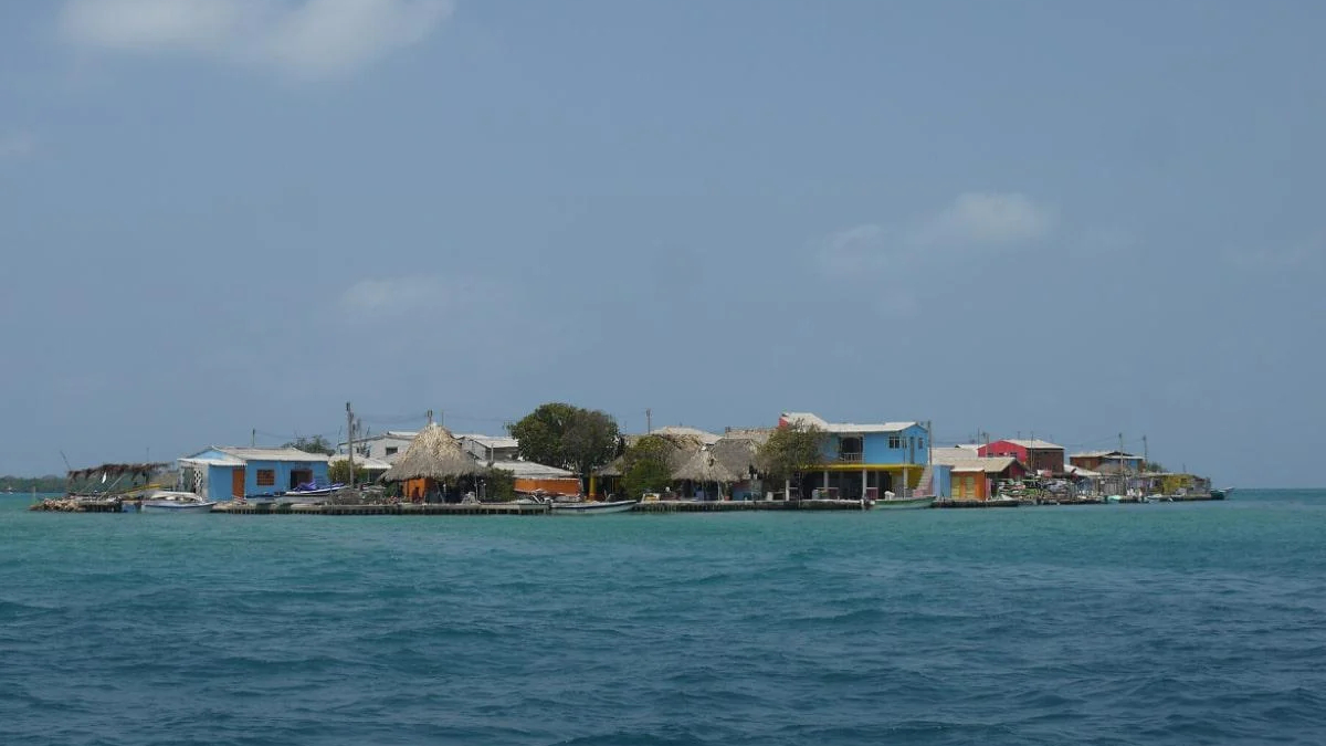 Small coastal village on a low-lying island with colorful houses and thatched roofs along a calm turquoise sea under a clear blue sky.