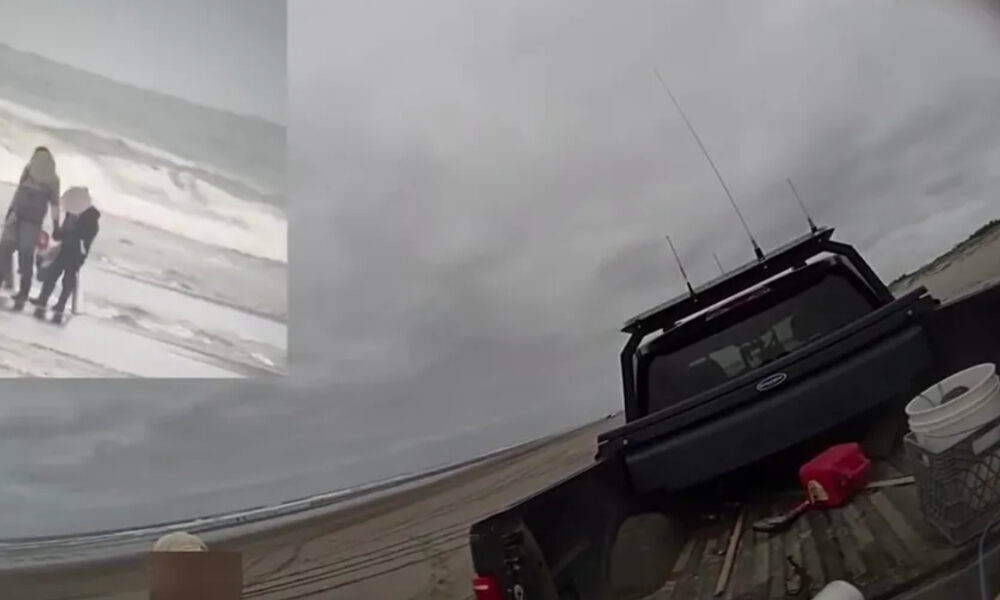 Three people stand on a windy, gray beach near incoming waves; a dark pickup truck sits on a wooden ramp by the shore.