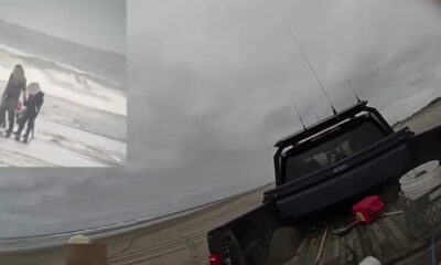 Three people stand on a windy, gray beach near incoming waves; a dark pickup truck sits on a wooden ramp by the shore.