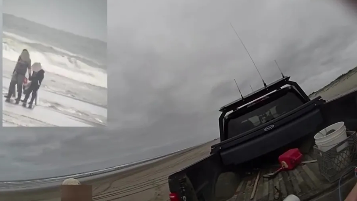 Three people stand on a windy, gray beach near incoming waves; a dark pickup truck sits on a wooden ramp by the shore.
