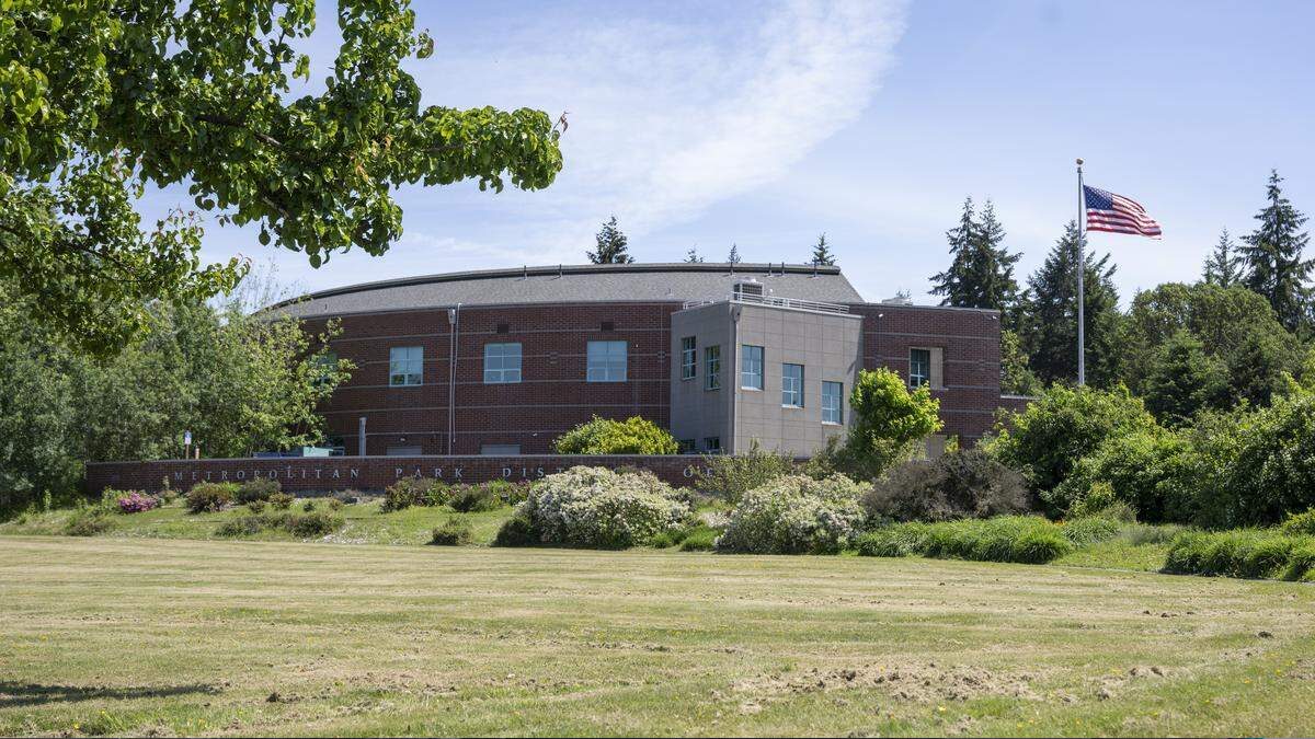 Municipal building with a brick exterior, a pale-gray entry tower, and an American flag on the right, surrounded by trees and a grassy lawn.