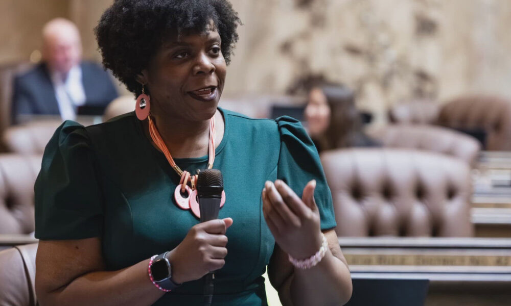 Woman in a teal dress speaks into a handheld microphone in a formal meeting room.
