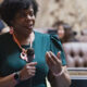 Woman in a teal dress speaks into a handheld microphone in a formal meeting room.
