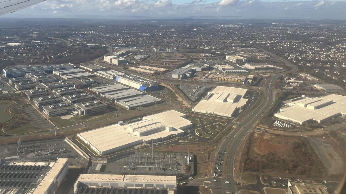 Aerial view of a large industrial park with white-roof warehouses, roads, and parking lots far from a residential area in the distance.
