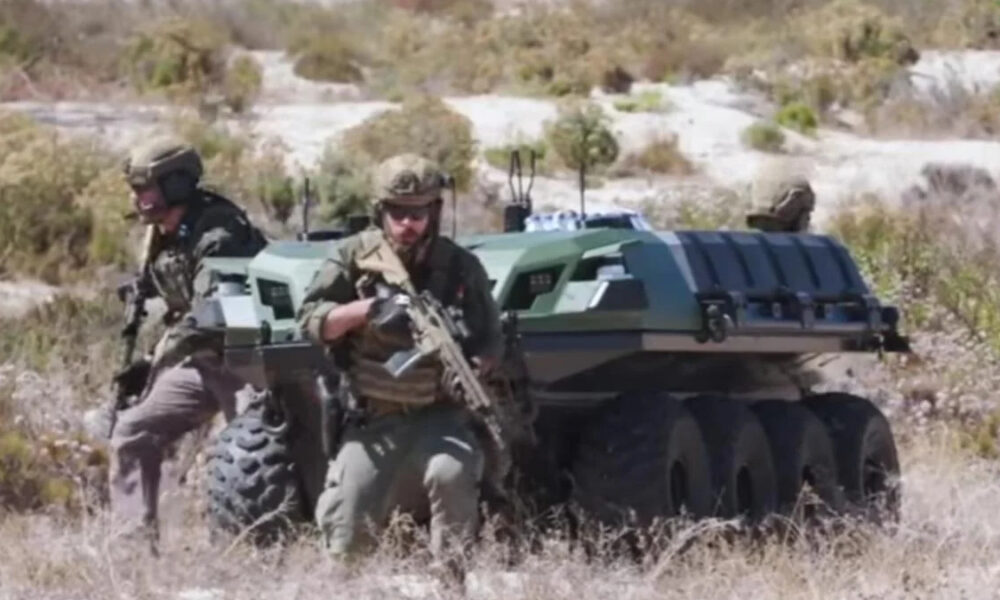 Soldiers in tactical gear with an armored, tracked vehicle in a desert landscape, advancing on foot and ready for action.