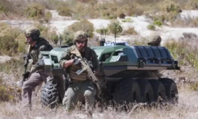 Soldiers in tactical gear with an armored, tracked vehicle in a desert landscape, advancing on foot and ready for action.