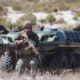 Soldiers in tactical gear with an armored, tracked vehicle in a desert landscape, advancing on foot and ready for action.