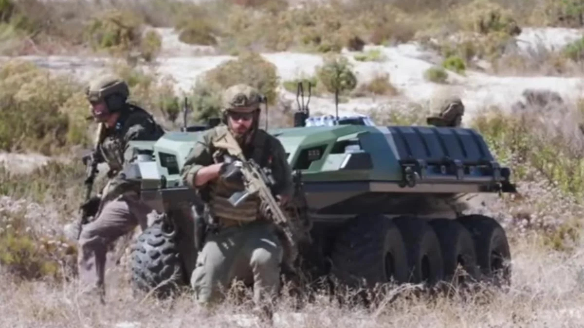 Soldiers in tactical gear with an armored, tracked vehicle in a desert landscape, advancing on foot and ready for action.