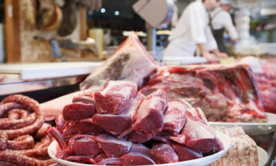 Piled raw red meat chops on a white platter at a market butcher stall, with sausages nearby.