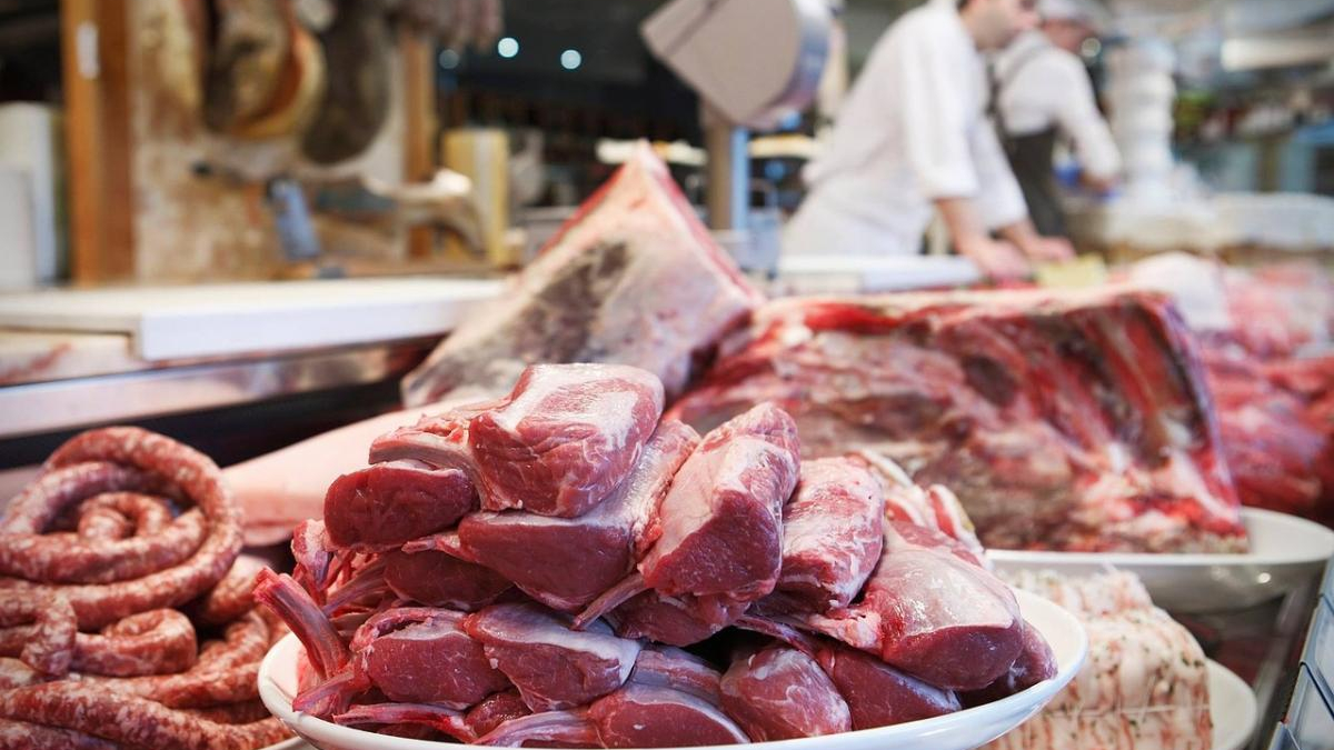 Piled raw red meat chops on a white platter at a market butcher stall, with sausages nearby.