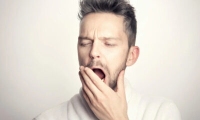 Man yawning with eyes closed, hand covering his mouth, wearing a white bathrobe/in a light-colored robe.