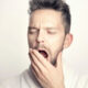 Man yawning with eyes closed, hand covering his mouth, wearing a white bathrobe/in a light-colored robe.