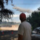 Man in a red cap stands on a dirt road watching a firefighting plane release smoke over a forest, with orange cones and a parked car nearby.