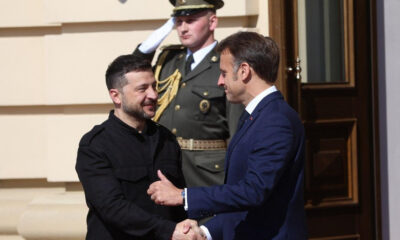 Two men shake hands outside a building as a uniformed guard salutes in the background.