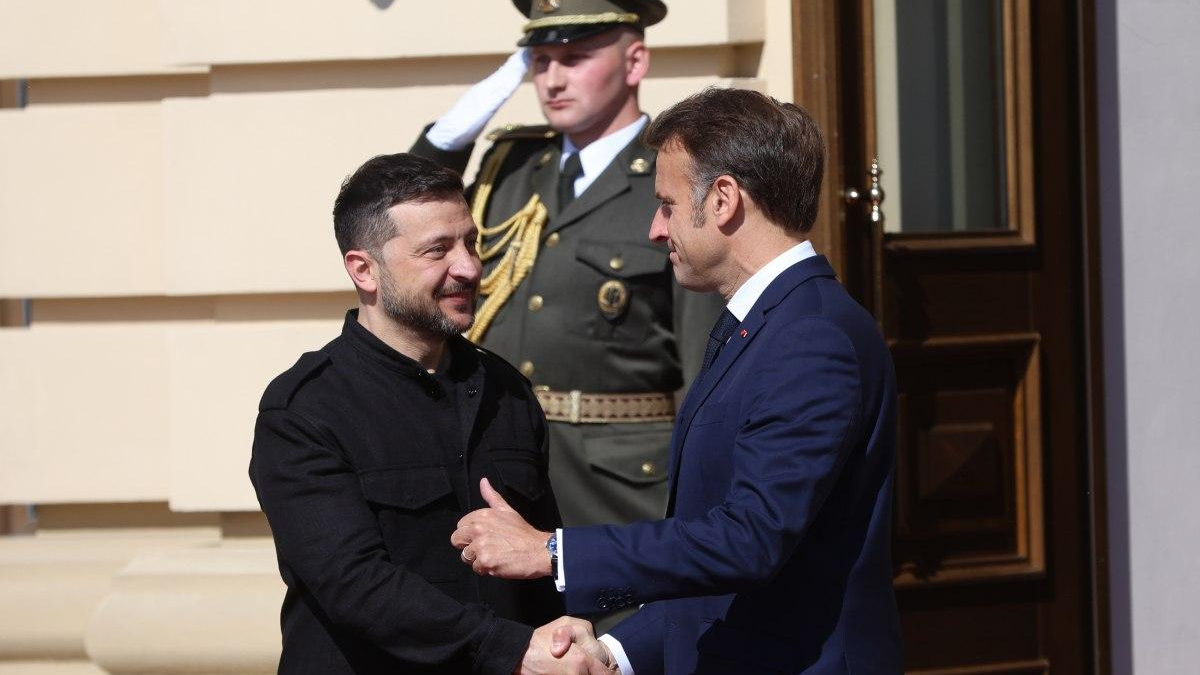 Two men shake hands outside a building as a uniformed guard salutes in the background.