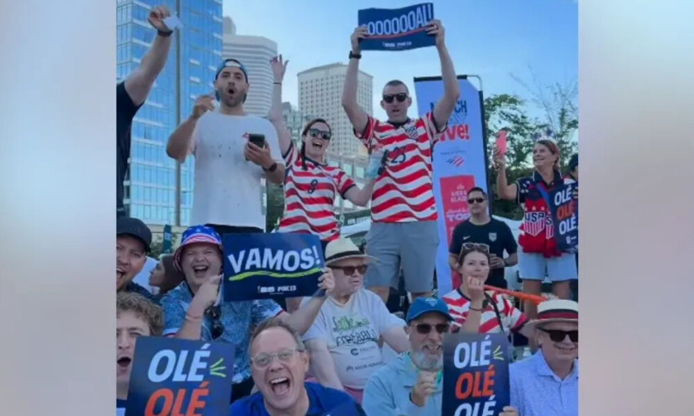Crowd of cheering spectators outdoors in a city, holding signs 'VAMOS!' and 'OLE OLE' with red‑white striped shirts.