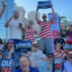 Crowd of cheering spectators outdoors in a city, holding signs 'VAMOS!' and 'OLE OLE' with red‑white striped shirts.