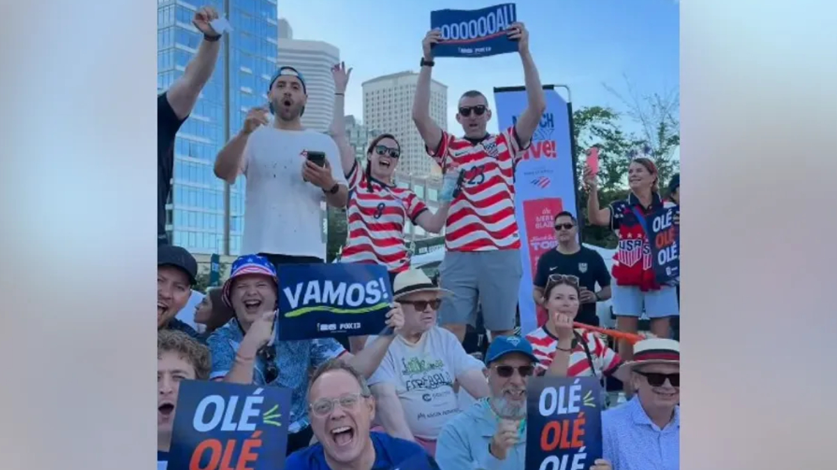 Crowd of cheering spectators outdoors in a city, holding signs 'VAMOS!' and 'OLE OLE' with red‑white striped shirts.