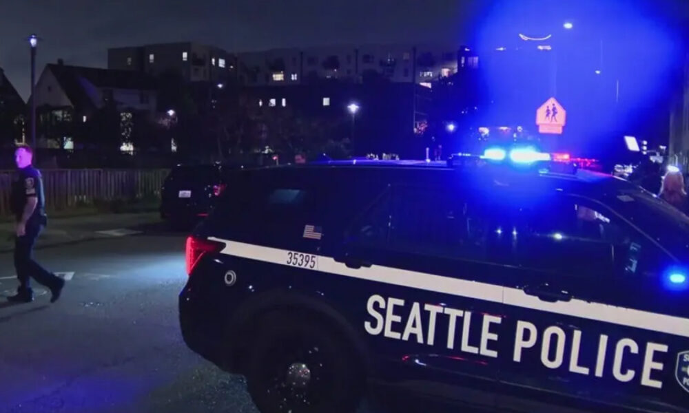 Night scene of a Seattle Police SUV with blue emergency lights flashing on a street, with pedestrians nearby.