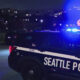 Night scene of a Seattle Police SUV with blue emergency lights flashing on a street, with pedestrians nearby.