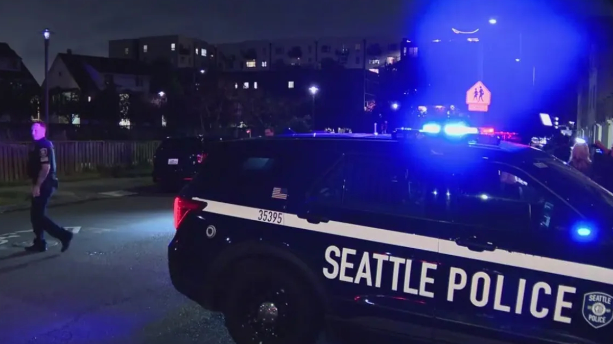 Night scene of a Seattle Police SUV with blue emergency lights flashing on a street, with pedestrians nearby.