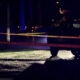 Nighttime street scene with police tape across the road, a dark SUV, and blue and red emergency lights.