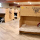 Dormitory-style shelter with wooden bunk beds along a hallway of a sunny, well-lit room.