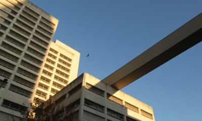 Two beige concrete buildings connected by a long elevated walkway against a clear blue sky; a bird flies nearby.