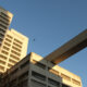 Two beige concrete buildings connected by a long elevated walkway against a clear blue sky; a bird flies nearby.