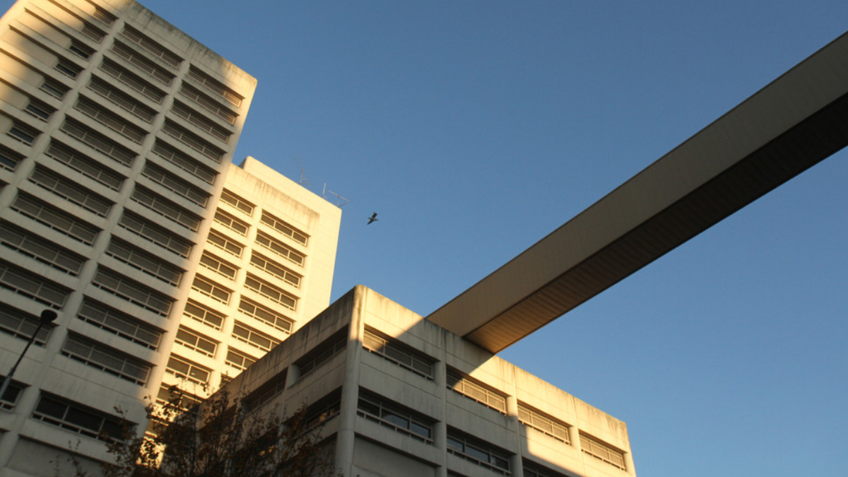 Two beige concrete buildings connected by a long elevated walkway against a clear blue sky; a bird flies nearby.