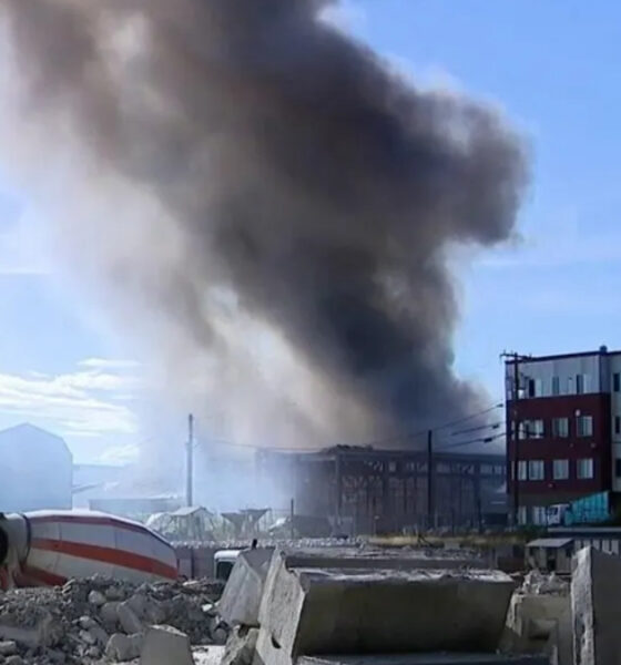 Large plume of dark smoke rises from a burning industrial area behind a rubble-filled construction site; a tilted cement mixer lies in the foreground.