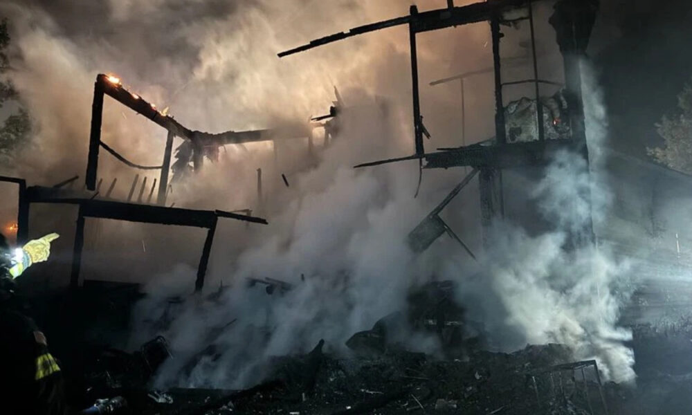 Night-time firefighting scene with a charred, collapsing wooden building and thick smoke; a firefighter with reflective gear points from the left side of the frame.