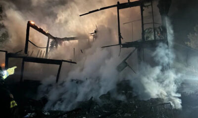 Night-time firefighting scene with a charred, collapsing wooden building and thick smoke; a firefighter with reflective gear points from the left side of the frame.