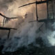 Night-time firefighting scene with a charred, collapsing wooden building and thick smoke; a firefighter with reflective gear points from the left side of the frame.