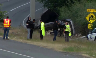 SUV flipped onto its side on the grassy roadside as responders and police stand nearby on a curve in the road.